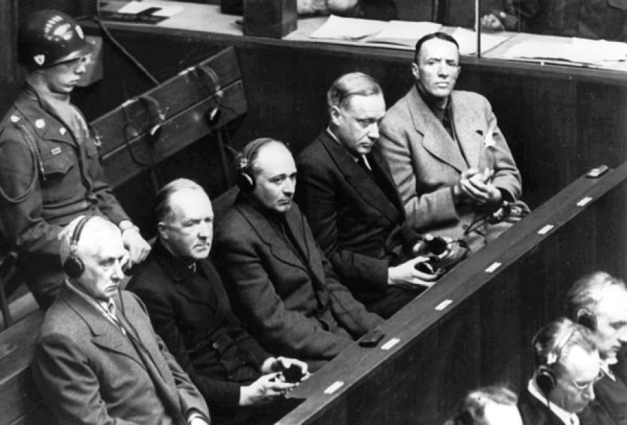 Image d'archives en noir et blanc avec cinq hommes assis dans les gradins au procès avec un garde derrière eux.