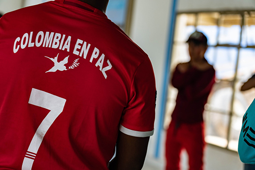 The back of a man wearing a soccer jersey that says "Colombia en paz" with the number 7, is in the foreground, with an out of focus second man in the background. 