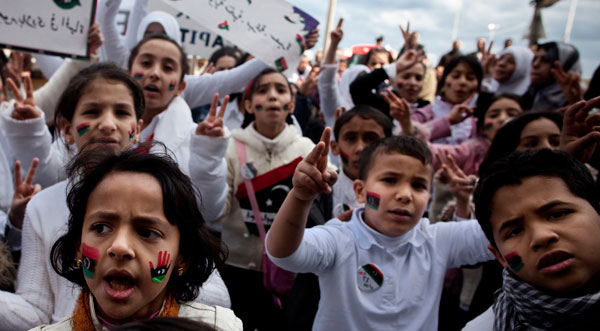 Children at a rally, holding up their fingers as peace signs with Libyan flags painted on their faces in red, black, and green. 