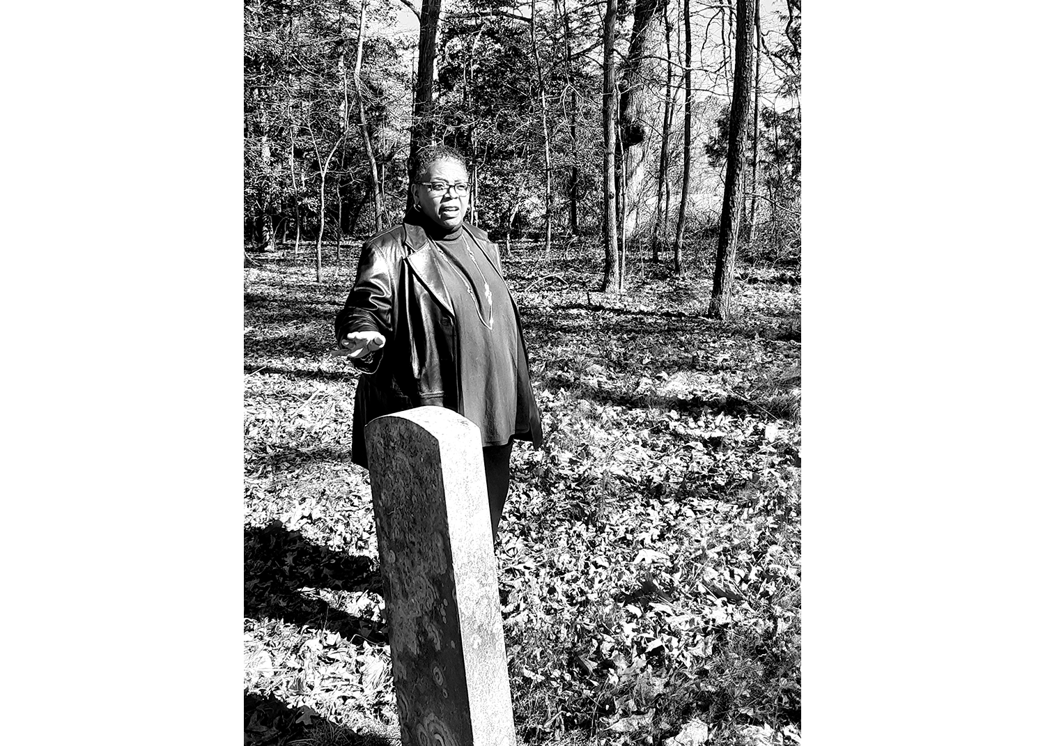 بالأسود والأبيض ، تقف امرأة سوداء تشير إلى شاهد قبر أمامها ، في مقبرة حولها العشب والأشجار.