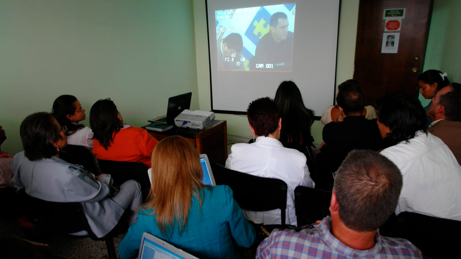 A small group of people seated in chairs in a small room watch a video screen on the wall.
