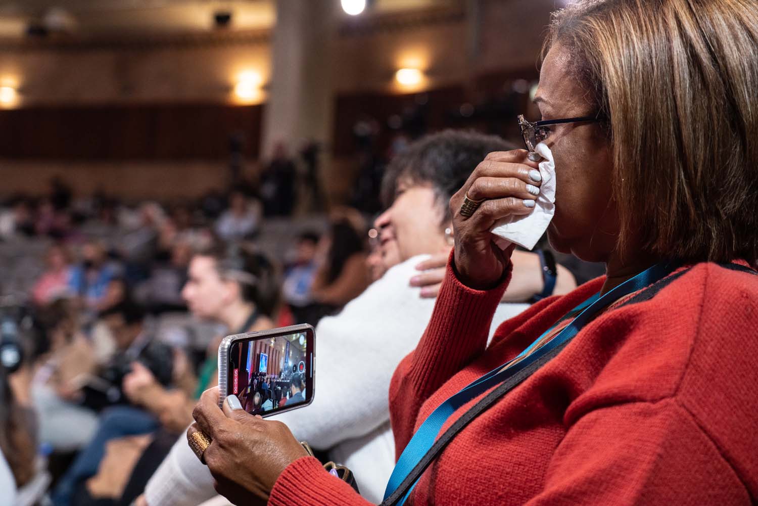 Une femme est assise dans le public, enregistrant l'audience sur son téléphone portable et essuyant ses larmes avec un mouchoir.