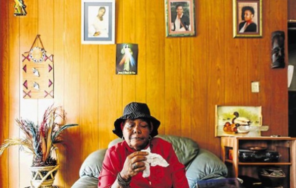 Ernestina Simelane sits holding a tissue, in front of a wood paneled wall with photos of her daughter hung behind her.