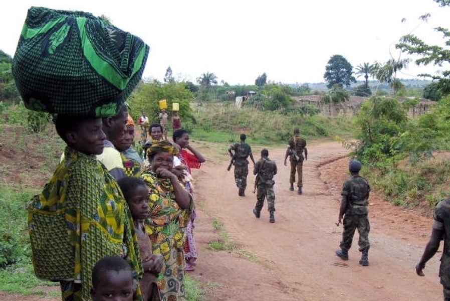 Woman and children stand on the left side, watching armed soldiers pass them on a dirt road.