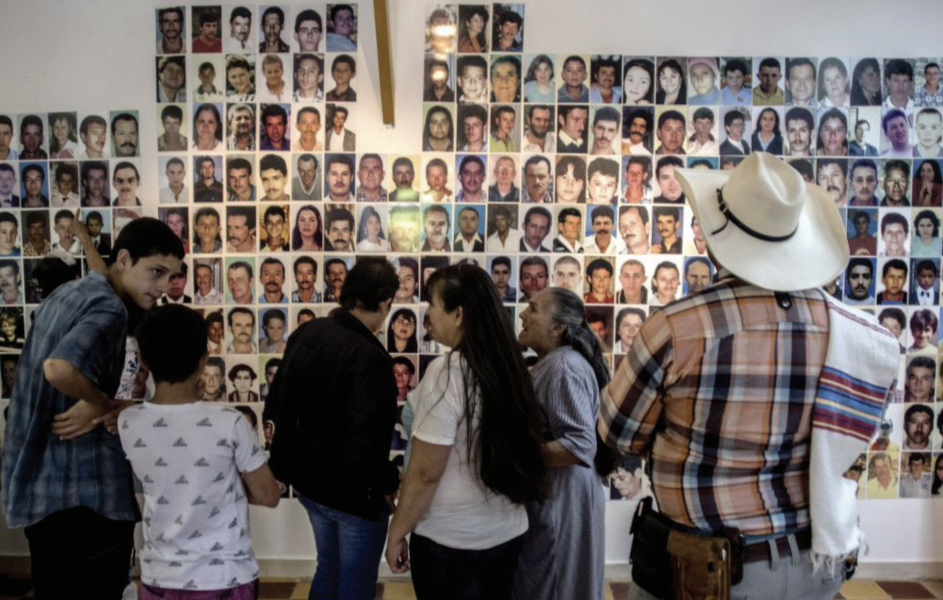 Children, women, and men look at a wall covered in faces of people. 