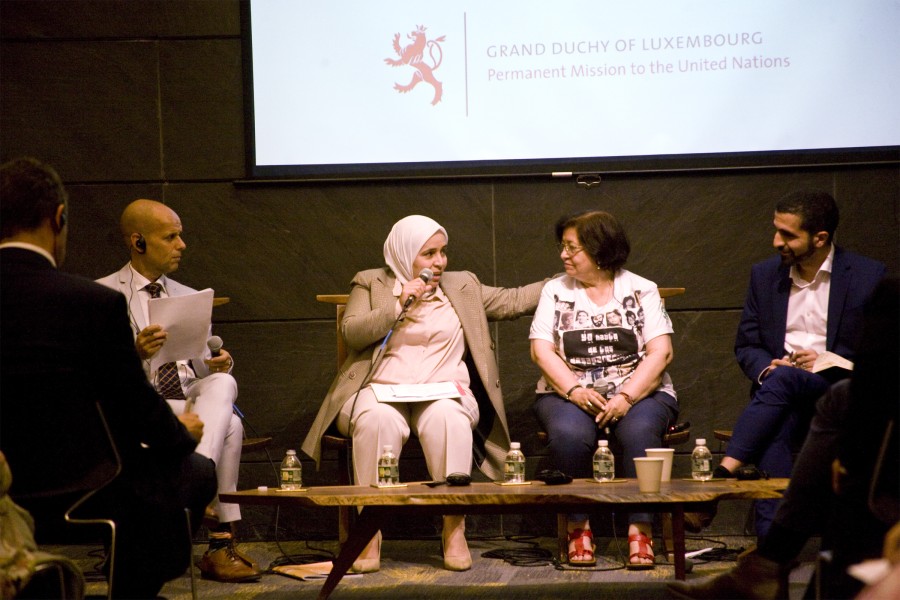 A panel of speakers sit in a darkened room, with two women in the center speaking to each other.