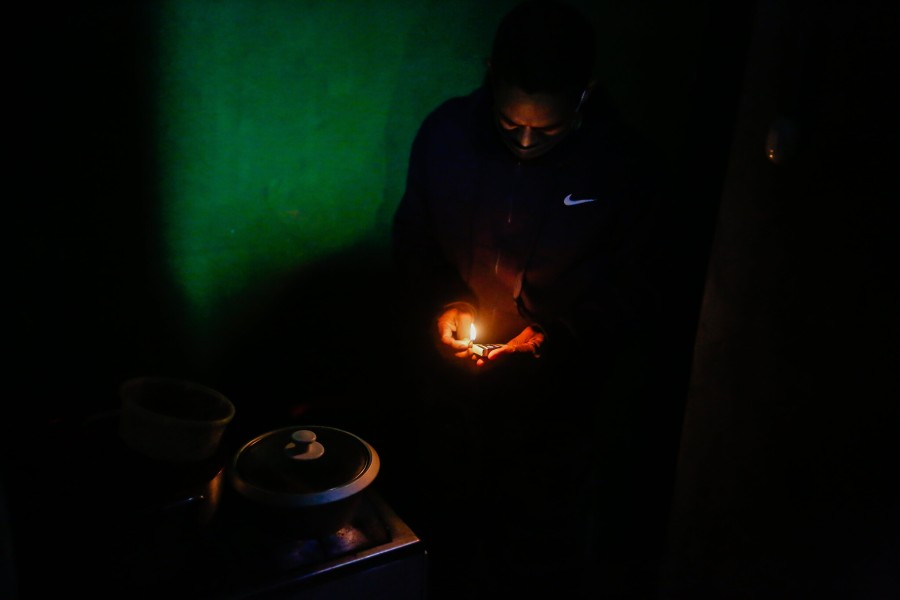 A man in a small dark room without electricity lights a match to start a hotplate where a pot sits.