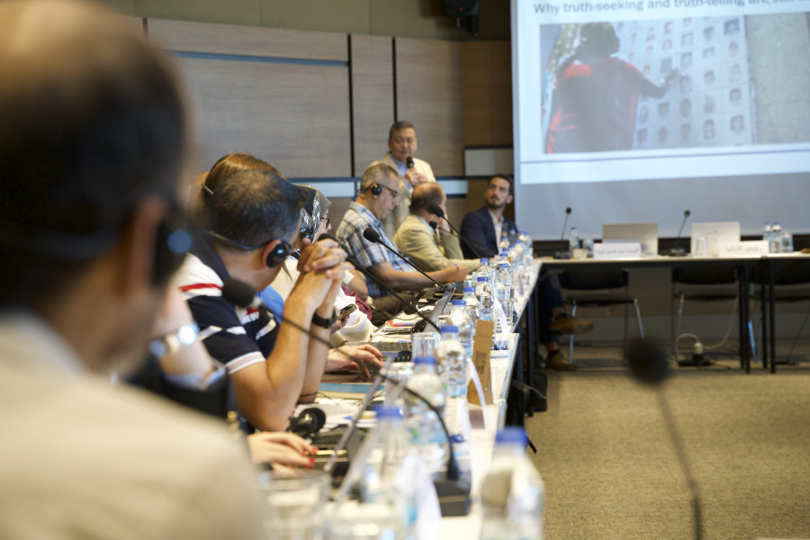 People sit along one side of a long table and look at a screen with the slide of a presentation