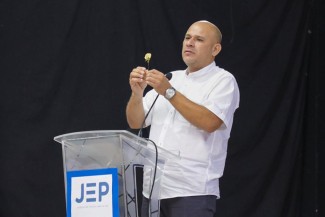 A man stands at a podium with a microphone, holding up a small white flower. 