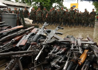 In the foreground is a large pile of military rifles. In the back stand a regiment of paramilitary fighters in army fatigues.