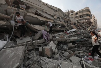 Two men and a women inspect the ruins of a destroyed building in an urban area in the daylight.
