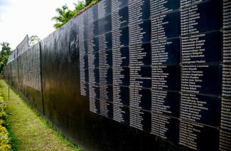 A black memorial wall lists names of victims. The wall is outdoors surrounded by green grass.