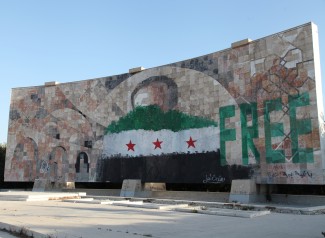 A large outdoor mural of a man's face is painted over with the Syrian revolution flag. Next to it, the English "Free" is painted in green. 