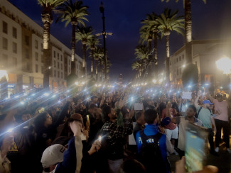 A crowd of people gather in the street in the evening; many hold up illuminated cell phones 