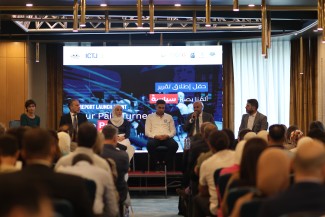 Six panelists sit in chairs on a stage facing a room full of people and the camera