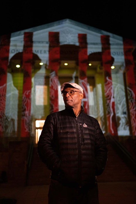 A man stands in front of a courthouse on which a digital artistic display is being projected.