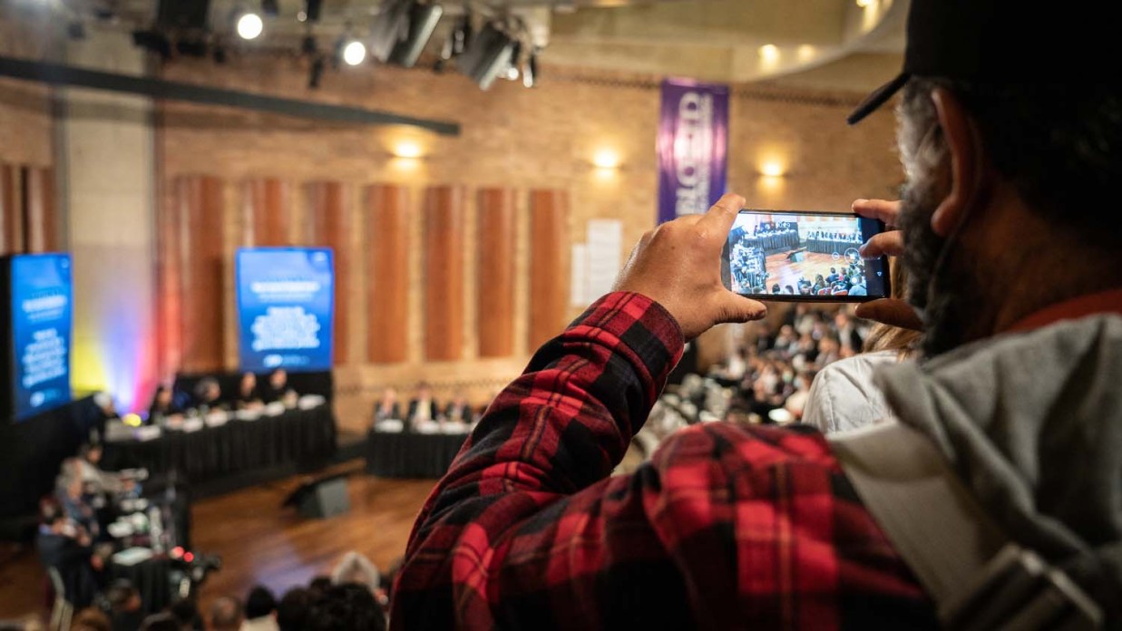 A man in the audience in the foreground uses a cell phone to record the hearing chamber in the background.