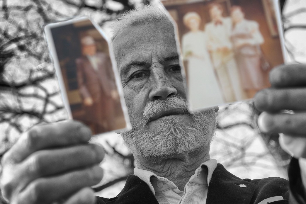 An old man holds two ripped halves of a photograph and looks out between them.