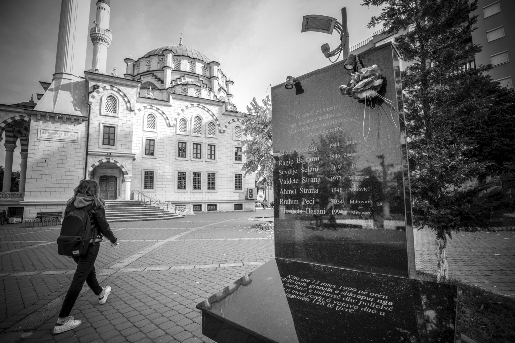 A youth with a backpack on walks away from the camera, around a monument in a city square.