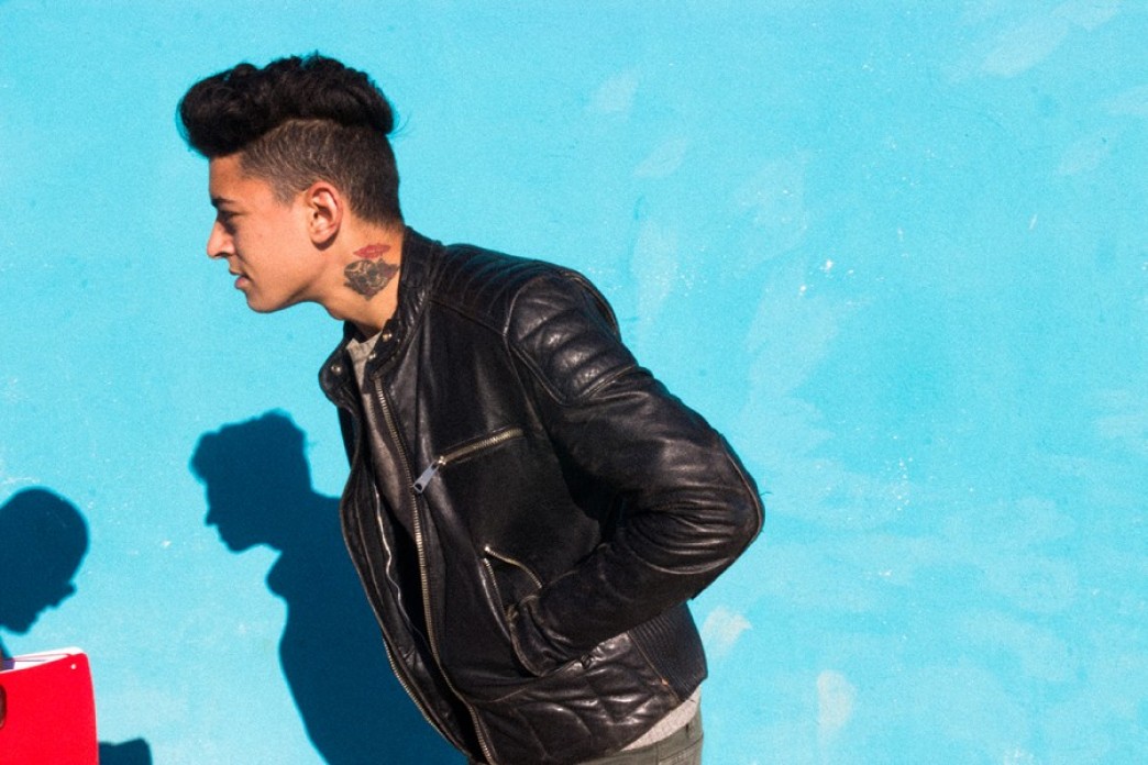 A man in a leather jacket stands in profile to the viewer with his hands in his jacket pockets, against a bright blue wall.