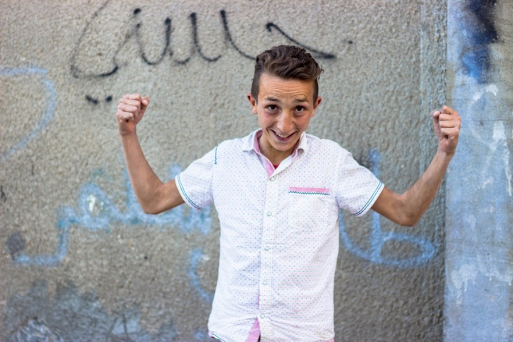 A boy stands with his arms flexed against a concrete wall with graffiti.