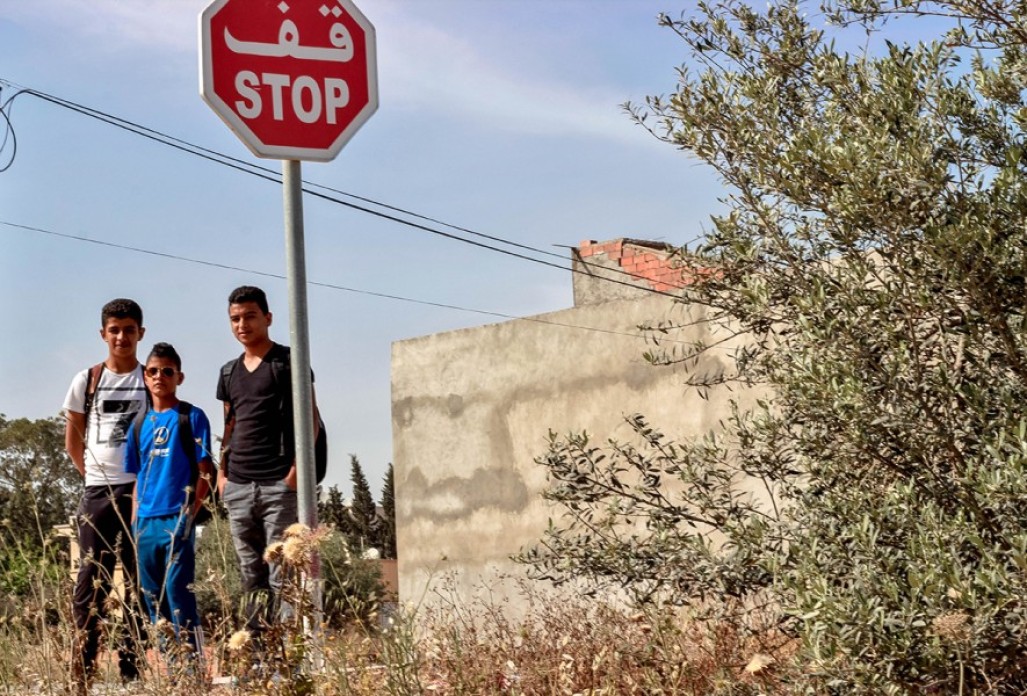 Three boys with backpacks on stand under a stop sign amidst dry shrubs and bushes.