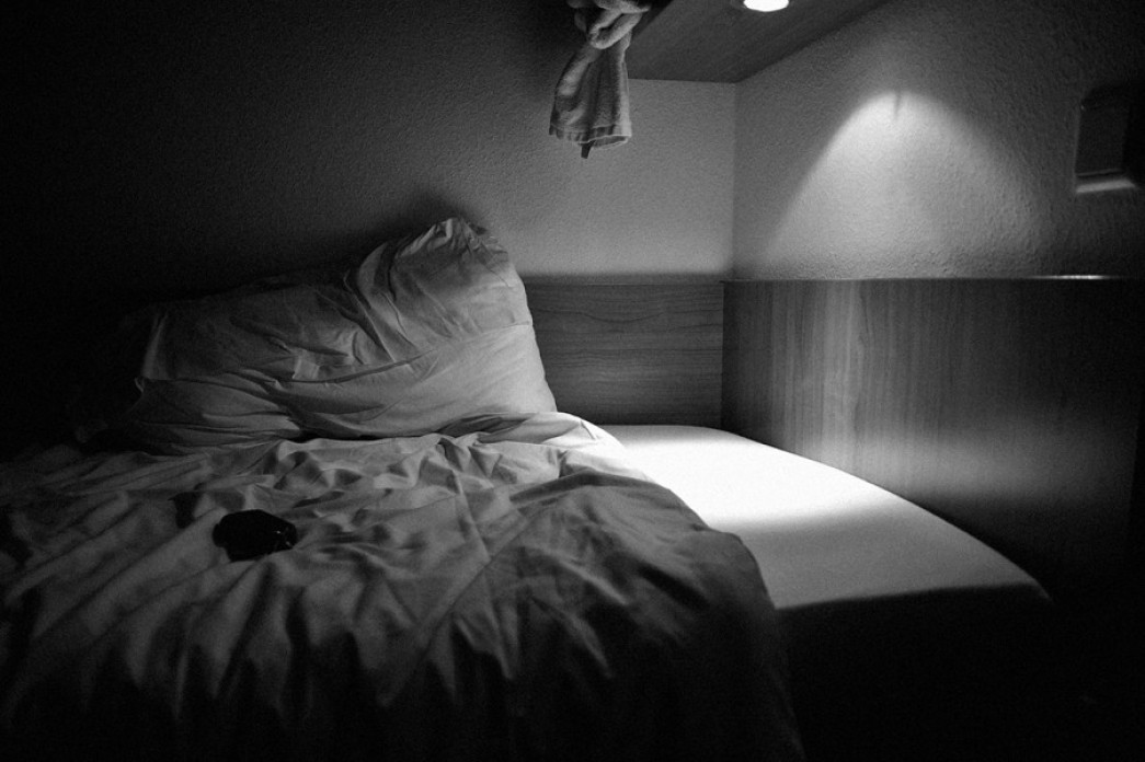 A photograph of a bed with a small part illuminated by a light and the rest in shadow, in black and white.