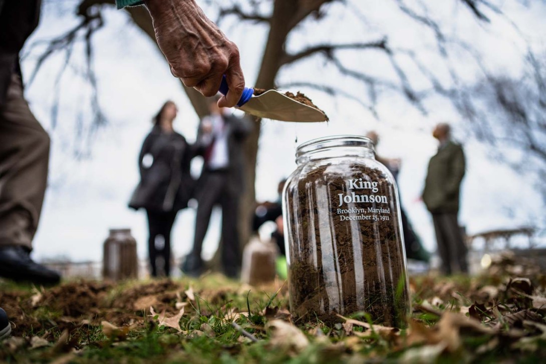 A clear glass jar with the name "King Johnson" with large white text, with "Brooklyn, Maryland, December 25, 1911," stands on the grass as a hand fills it with dirt.  