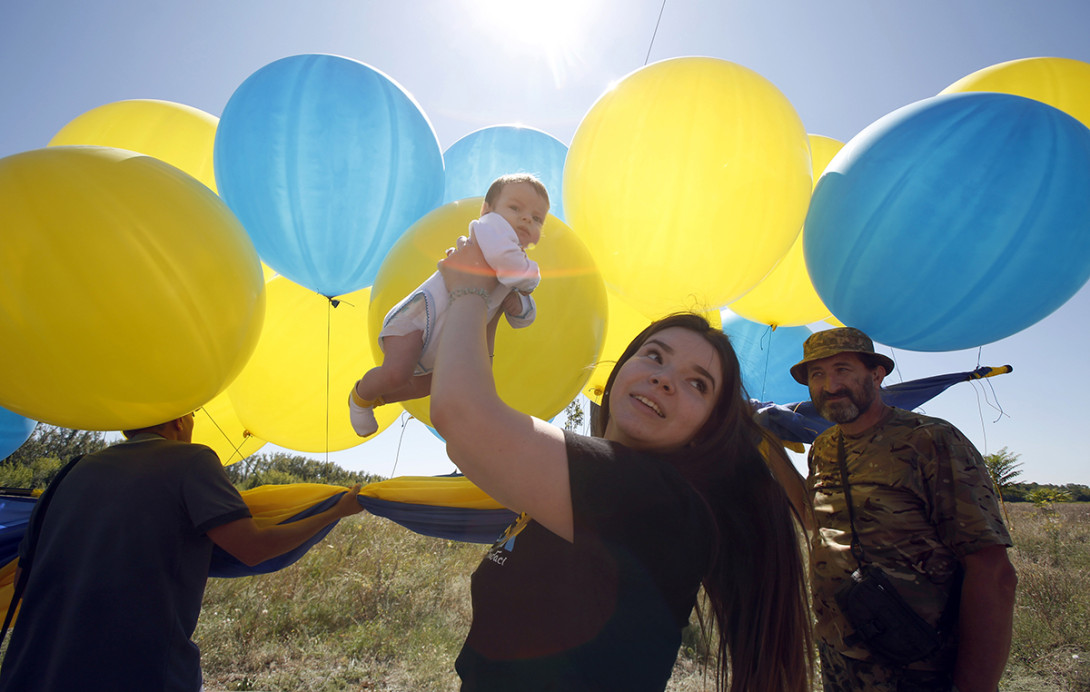 A women holds up an infant in the air on sunny day outdoors, surrounded by yellow and blue balloons